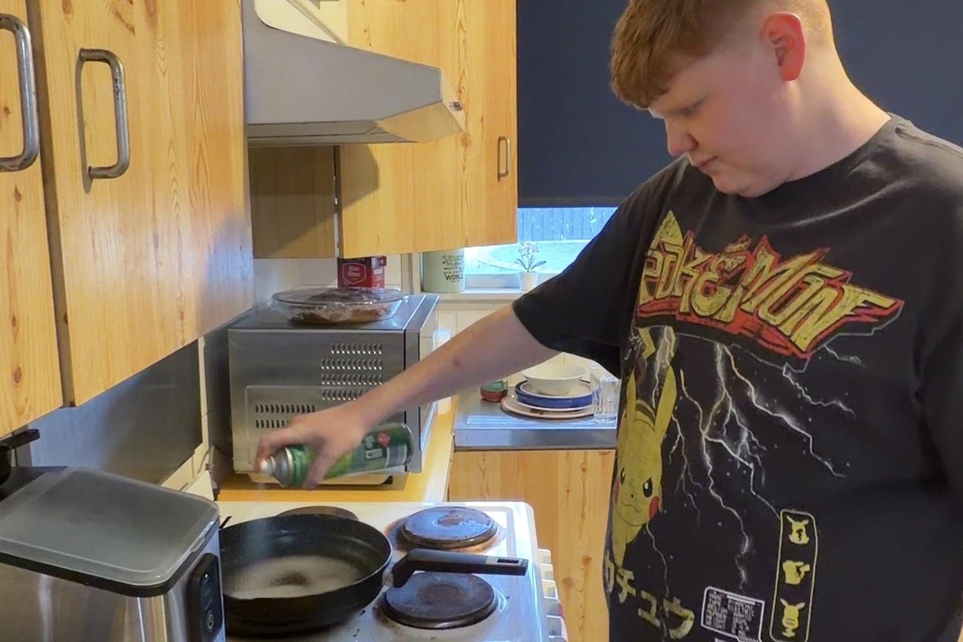 A young adult cooking, spraying oil into a frying pan on the stovetop.