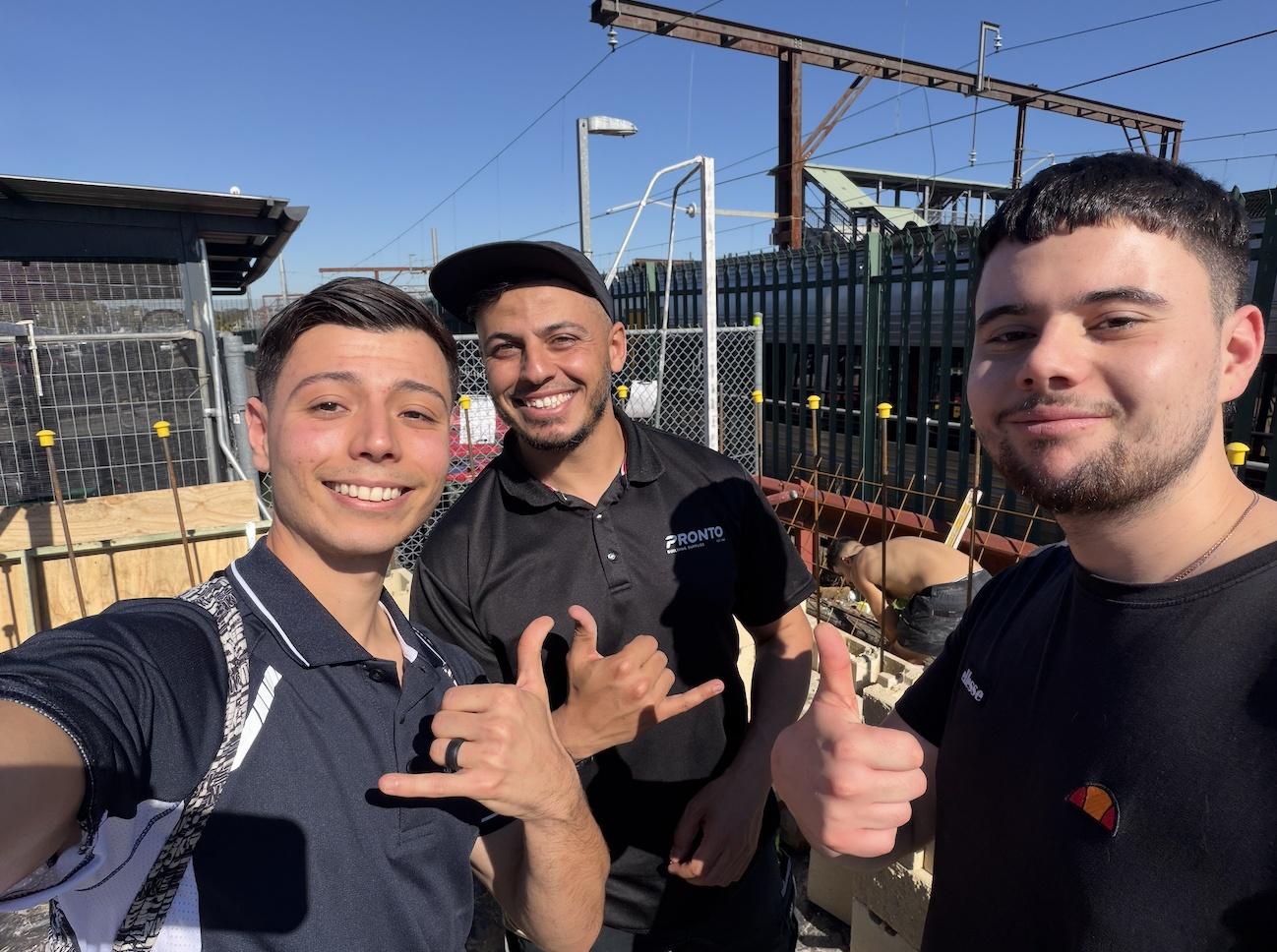 Three young men smiling at an outdoor building site, one giving a thumbs up and one a hang loose sign.